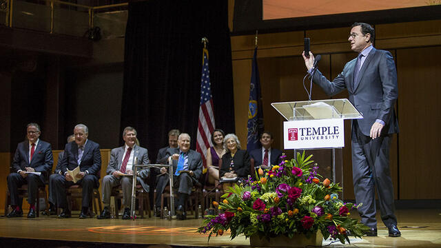 Bob Saget holding up his cell phone as he stands at a podium.