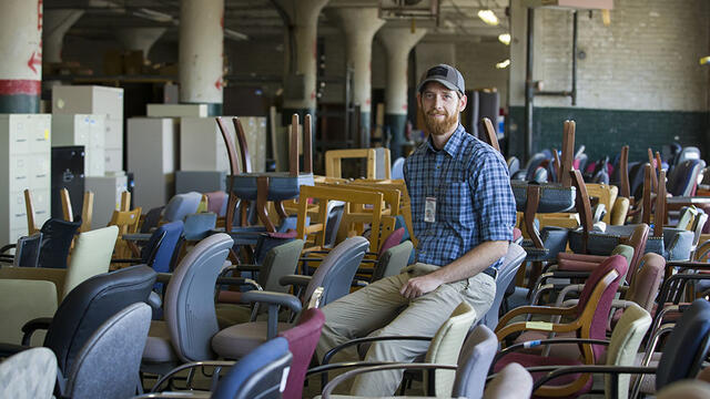 A man sitting amid cabinets, desks, chairs and other items in a warehouse.