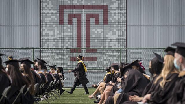 a graduate in a robe and cap walks in front of a large Temple T