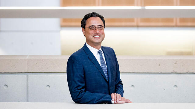 A man with dark hair and glasses wears a blue suit and smiles at a camera.