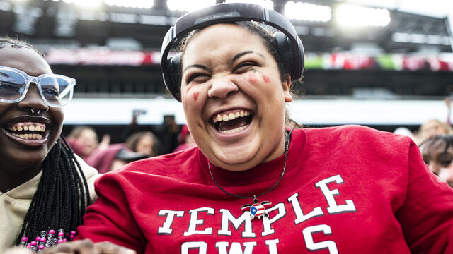 Image of a college student smiling, wearing a cherry and white shirt, seated at a Temple University homecoming event.