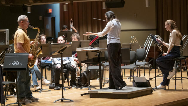Image of Matthew Levy soloing with the wind symphony. Patricia Cornett conducts while Emma O'Halloran observes.