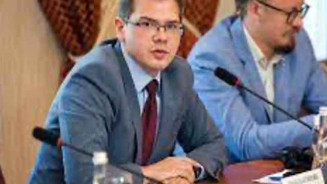 image of Ivan wearing glasses, in a suit sitting at a table speaking into a microphone