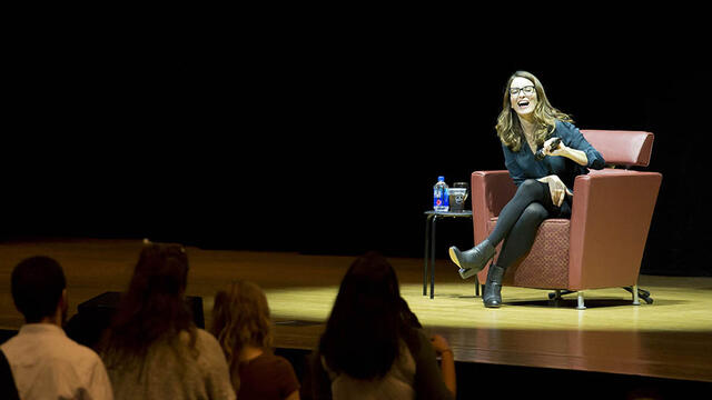 Tina Fey sitting on stage while holding a microphone and laughing.
