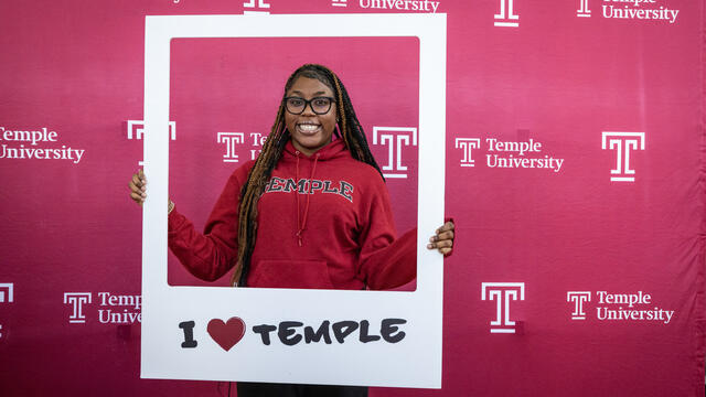 A student wearing a Temple hoodie poses in a frame that says "I heart Temple"
