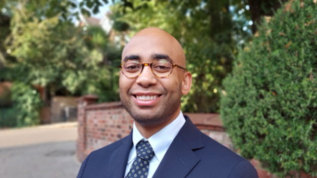 Man with wire glasses wearing blue suit, light blue shirt and blue tie in a park 