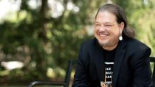 image of Jeremy Johnson sitting on a bench wearing a black blazer, jeans and is smiling looking into the distance