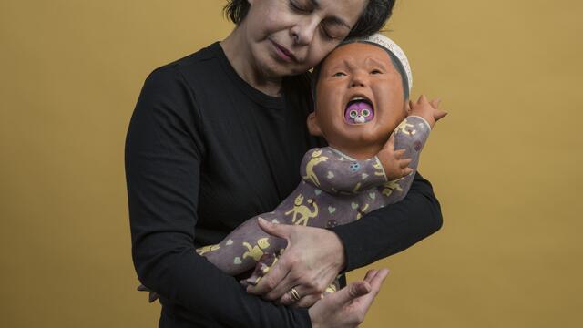 Woman holding Baby clay sculpture that appears to be crying has purple round object in mouth