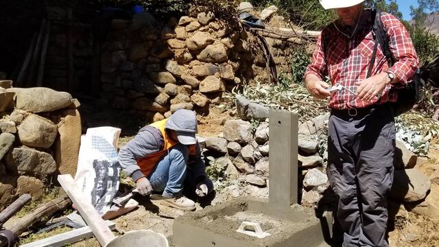 Two people working at an outdoor stone construction site, placing and measuring materials near a partially built stone structure, with buckets and tools in the foreground.