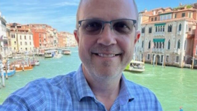 image of Brad standing in Venice wearing glasses and a blue shirt smiling at the camera
