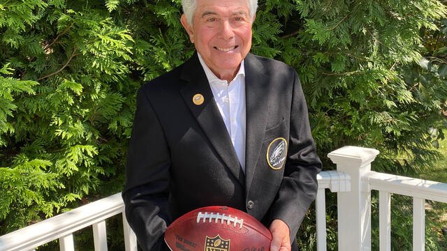 Merrill Reese holding a football and wearing his Eagles Hall of Fame jacket outside.