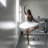 A Black woman in white tutu practicing ballet at a barre in a dance studio.