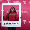 Student wearing a Temple hoodie poses in a frame that says "I heart Temple"