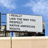 A billboard sits atop a building in a blue sky. In black letters on white background, the billboard reads "I REALLY LIKE THE WAY YOU RESPECT NATIVE AMERICAN RIGHTS."